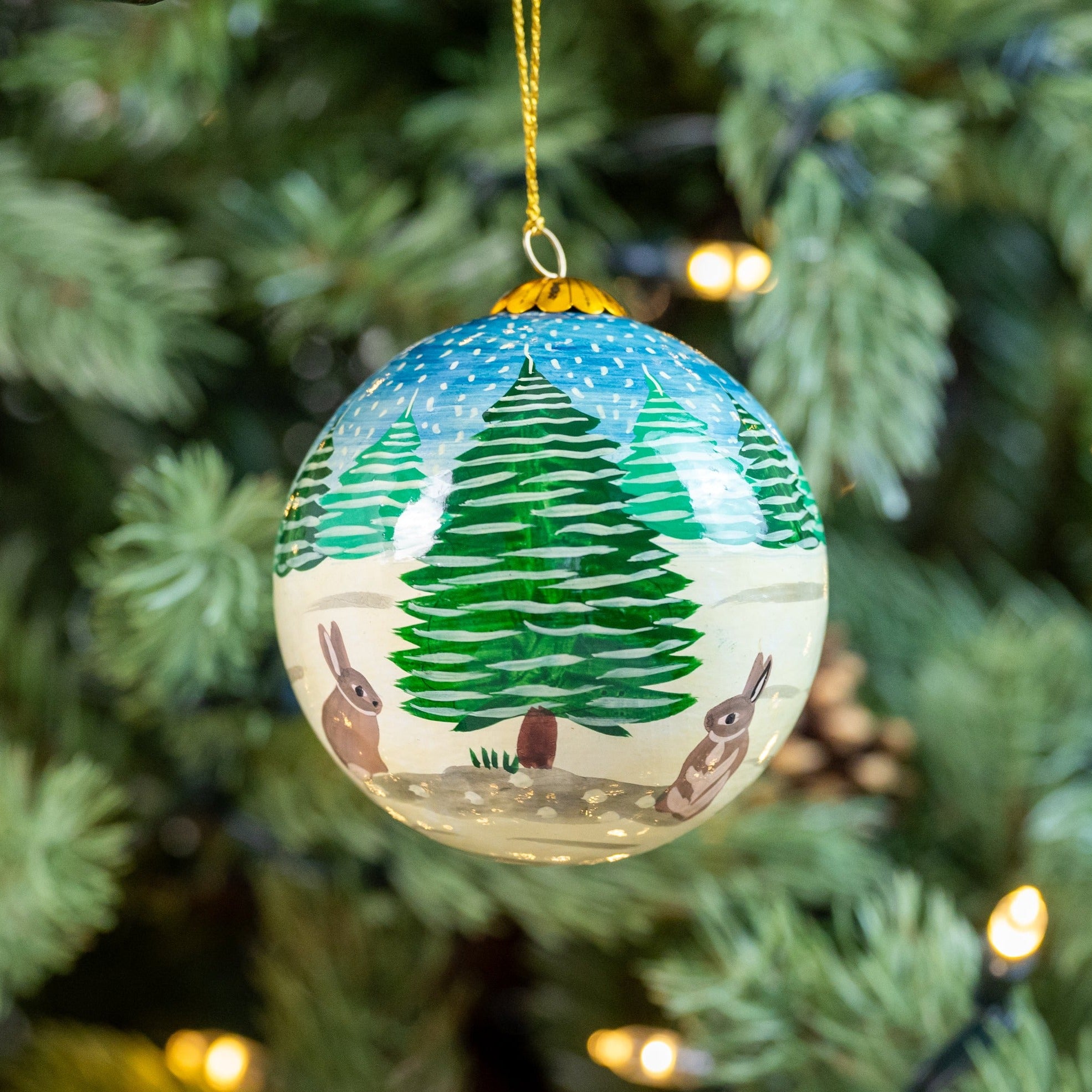 A hand-painted Christmas bauble with a scene of rabbits in a snowy landscape, hanging on a tree with lights and decorations.
