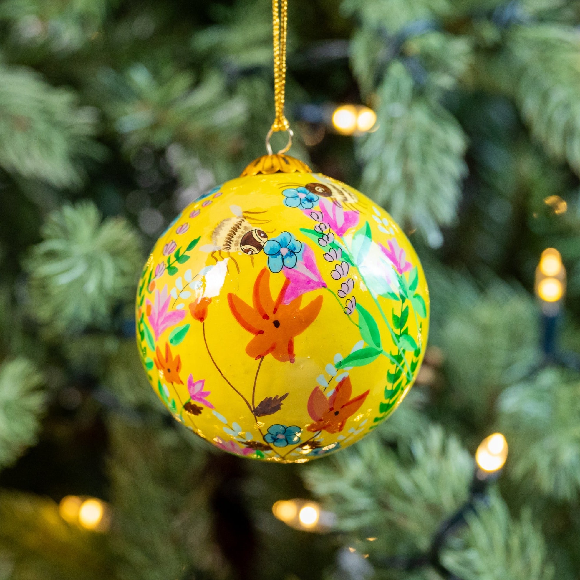 Hand painted yellow Christmas bauble with a floral and bee design, hanging on a tree with lights and needles in the background.