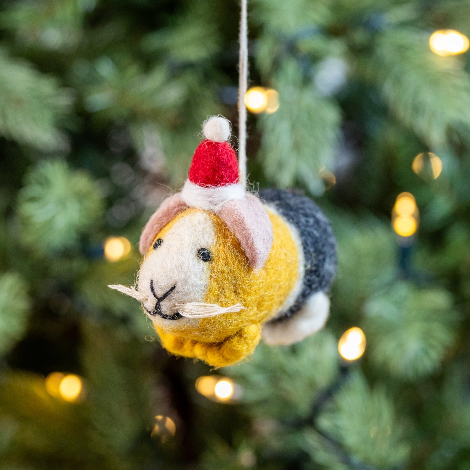 Handmade wool felt decoration of a guinea pig wearing a Santa hat, hanging on a Christmas tree.