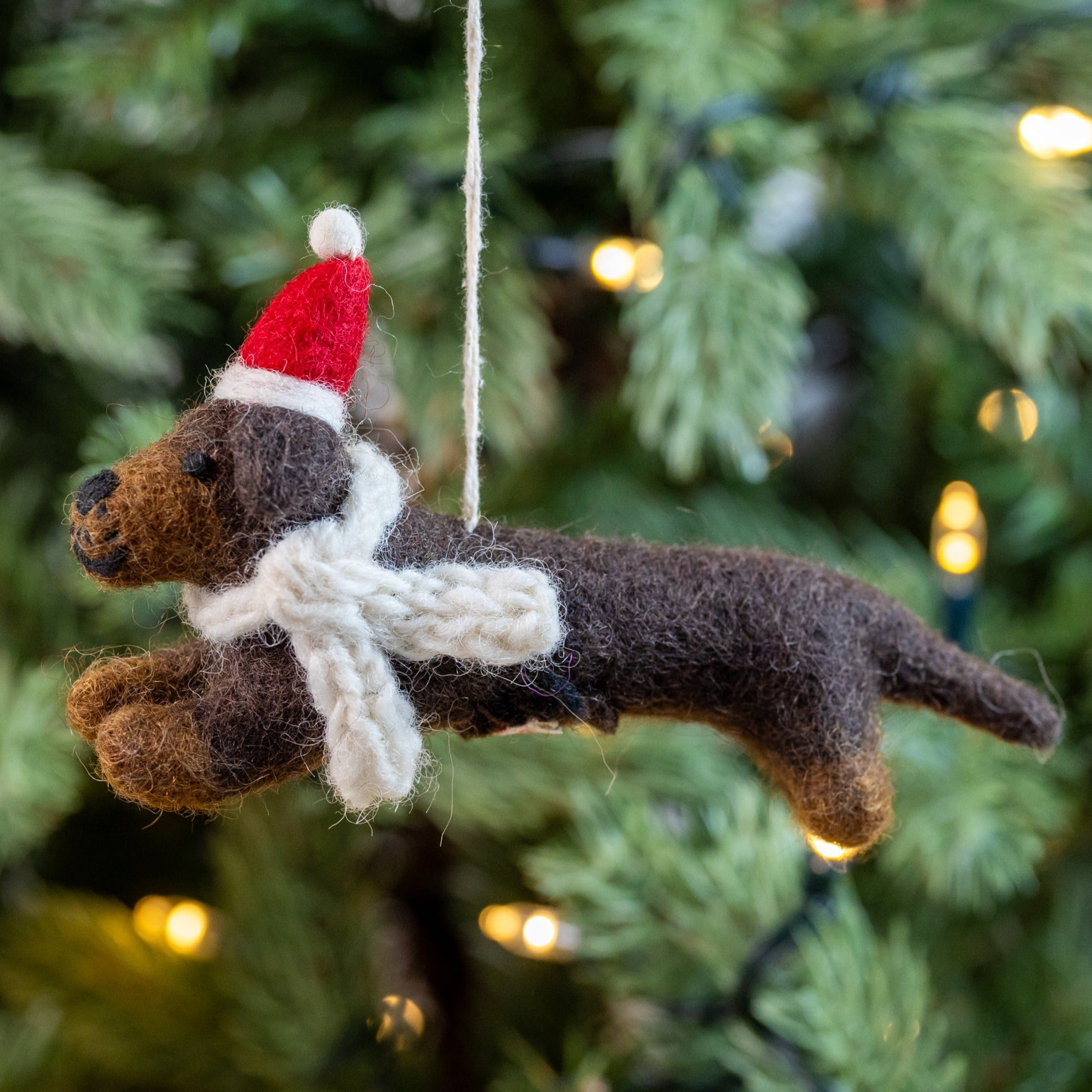 A handmade felt decoration of a sausage dog wearing a hat and scarf, designed to be hung as a Christmas decoration.