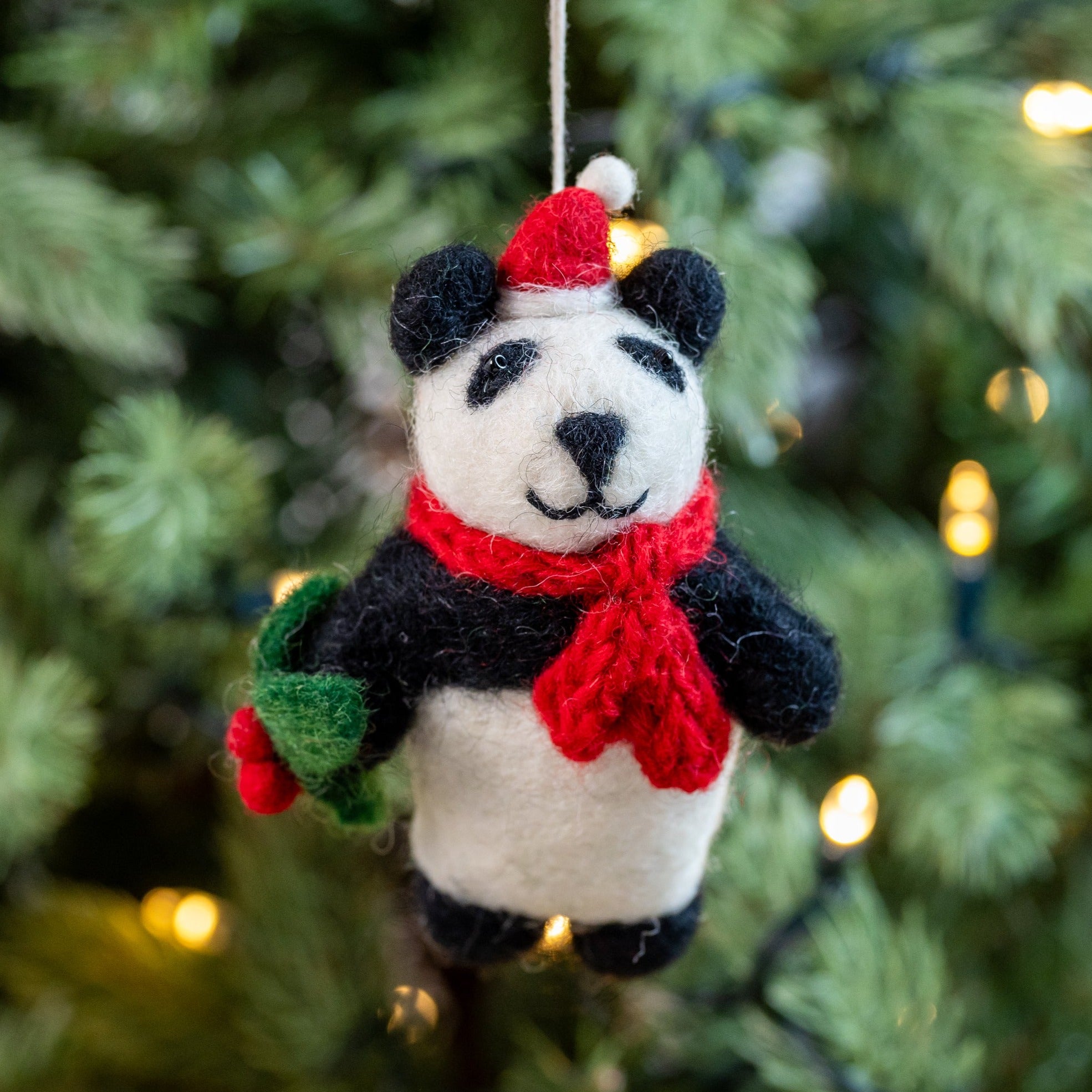 A handmade felt panda decoration with a red and green scarf, hanging from a Christmas tree with blurred lights in the background.