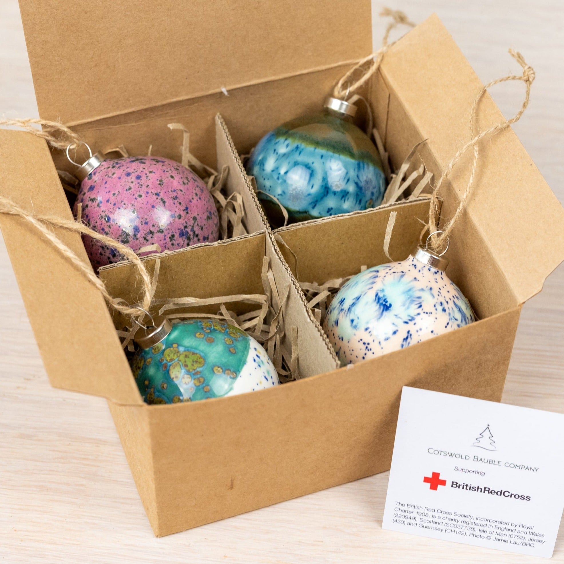 A set of four handmade ceramic Christmas baubles in pink, blue, turquoise, and cream colors, displayed inside an open cardboard box with each bauble threaded with biodegradable jute twine. The baubles have a diamond shape and the packaging is made from recycled materials.