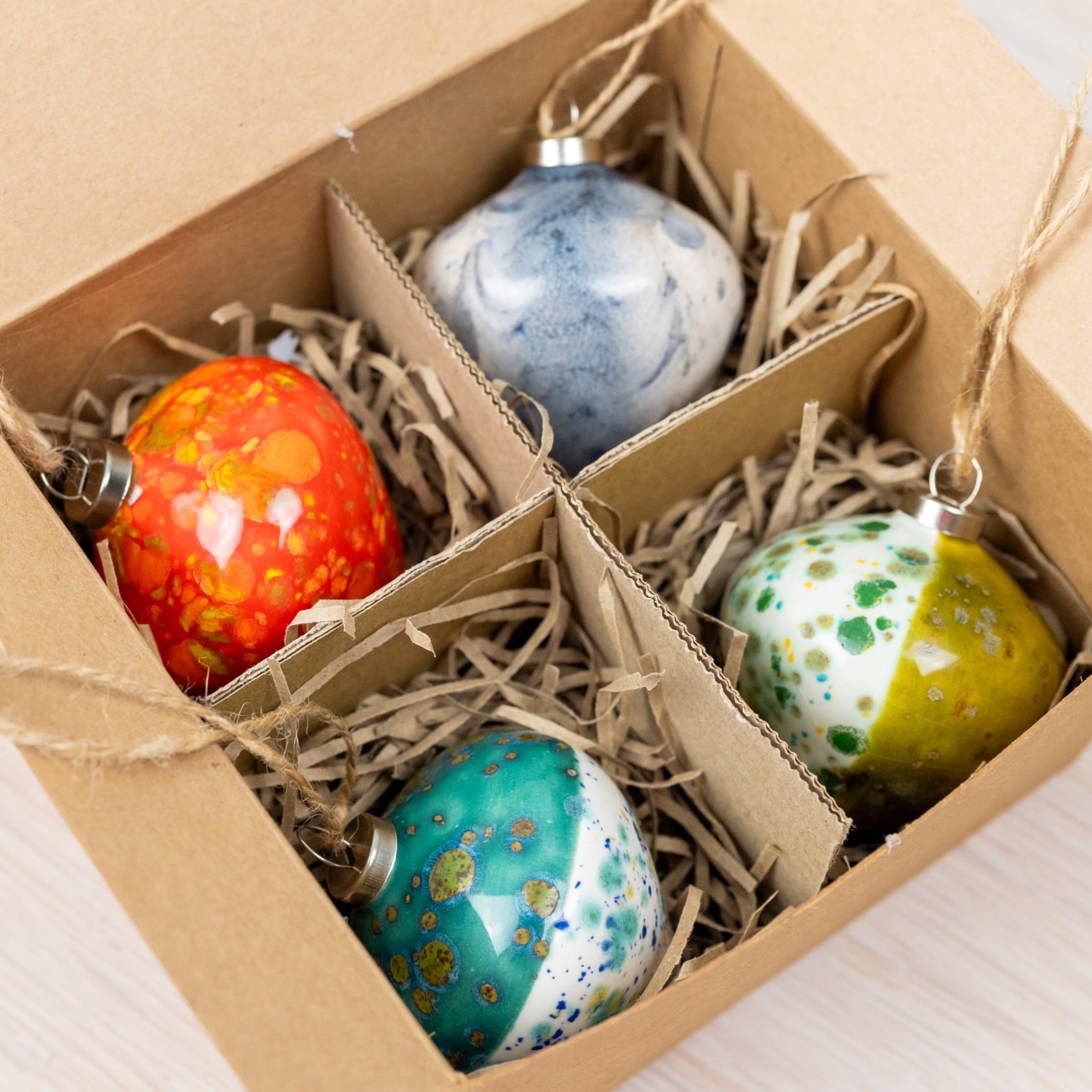 A set of four handmade ceramic Christmas baubles in green, red, grey, and turquoise diamond shapes, displayed in an open box with each bauble threaded with biodegradable jute twine.