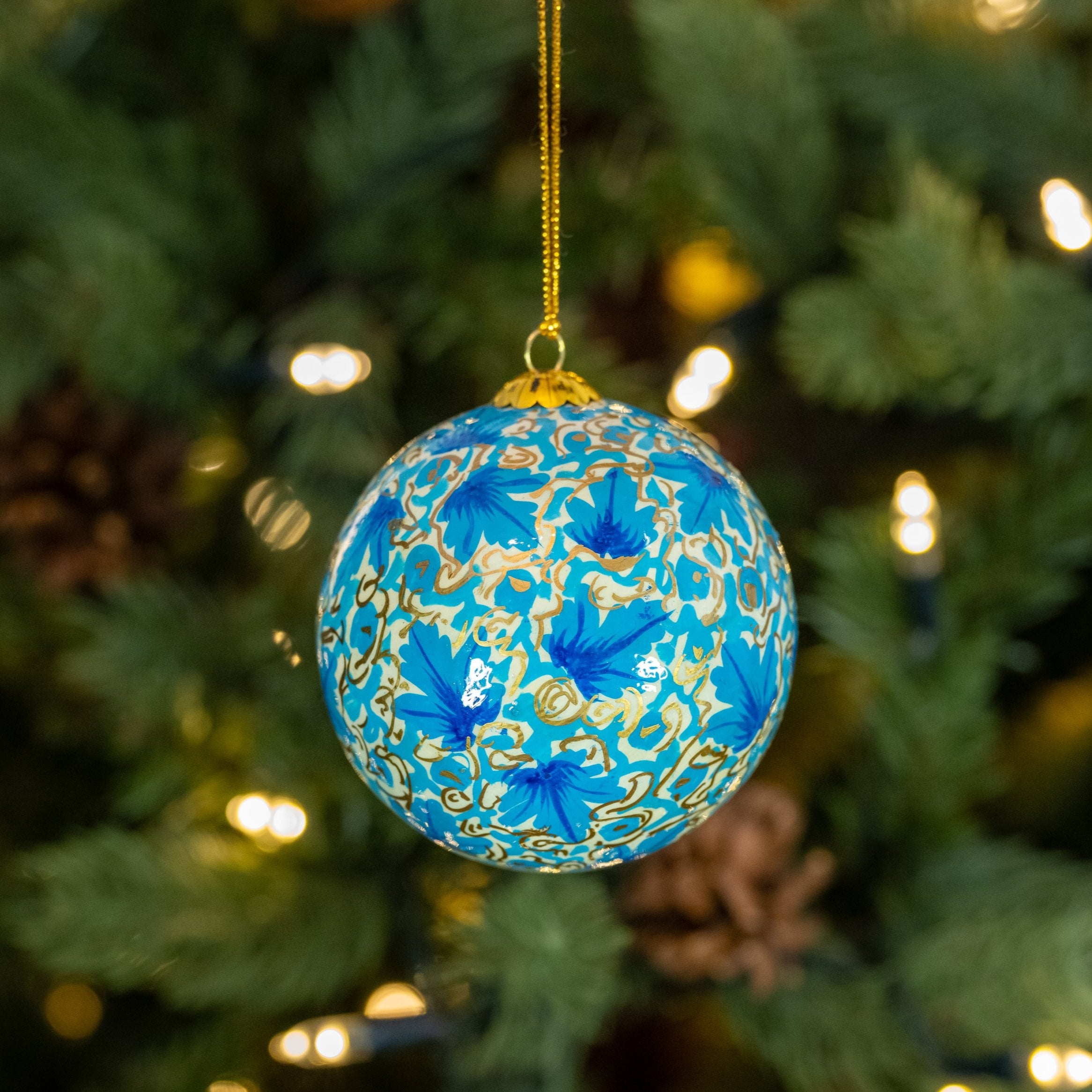 A hand painted blue Christmas bauble with a floral design, hanging on a Christmas tree.