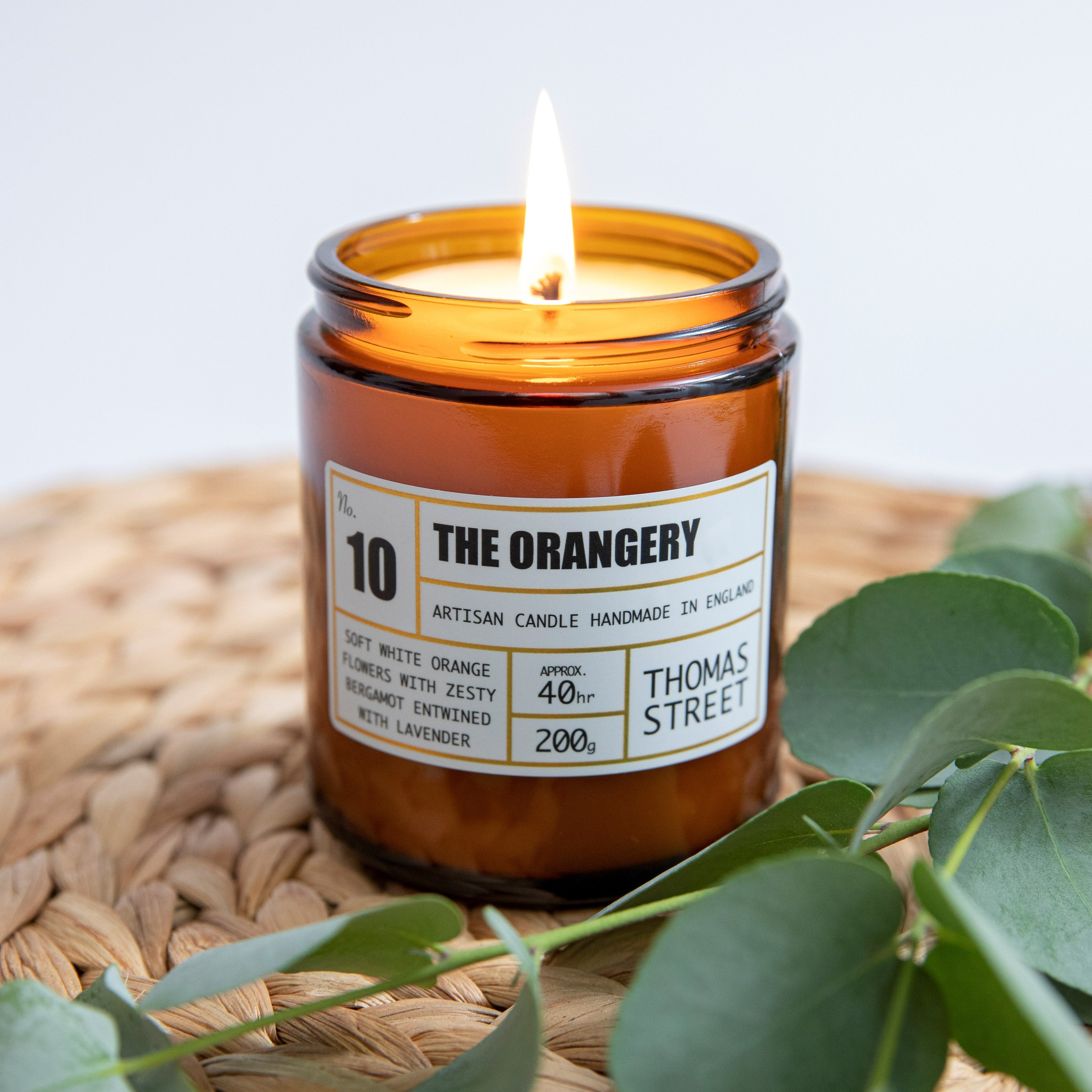 A scented candle with a lit wick, titled 'The Orangery' by Thomas Street, on a wicker surface with green leaves and a label indicating 40 hours burn time, and 200g weight.