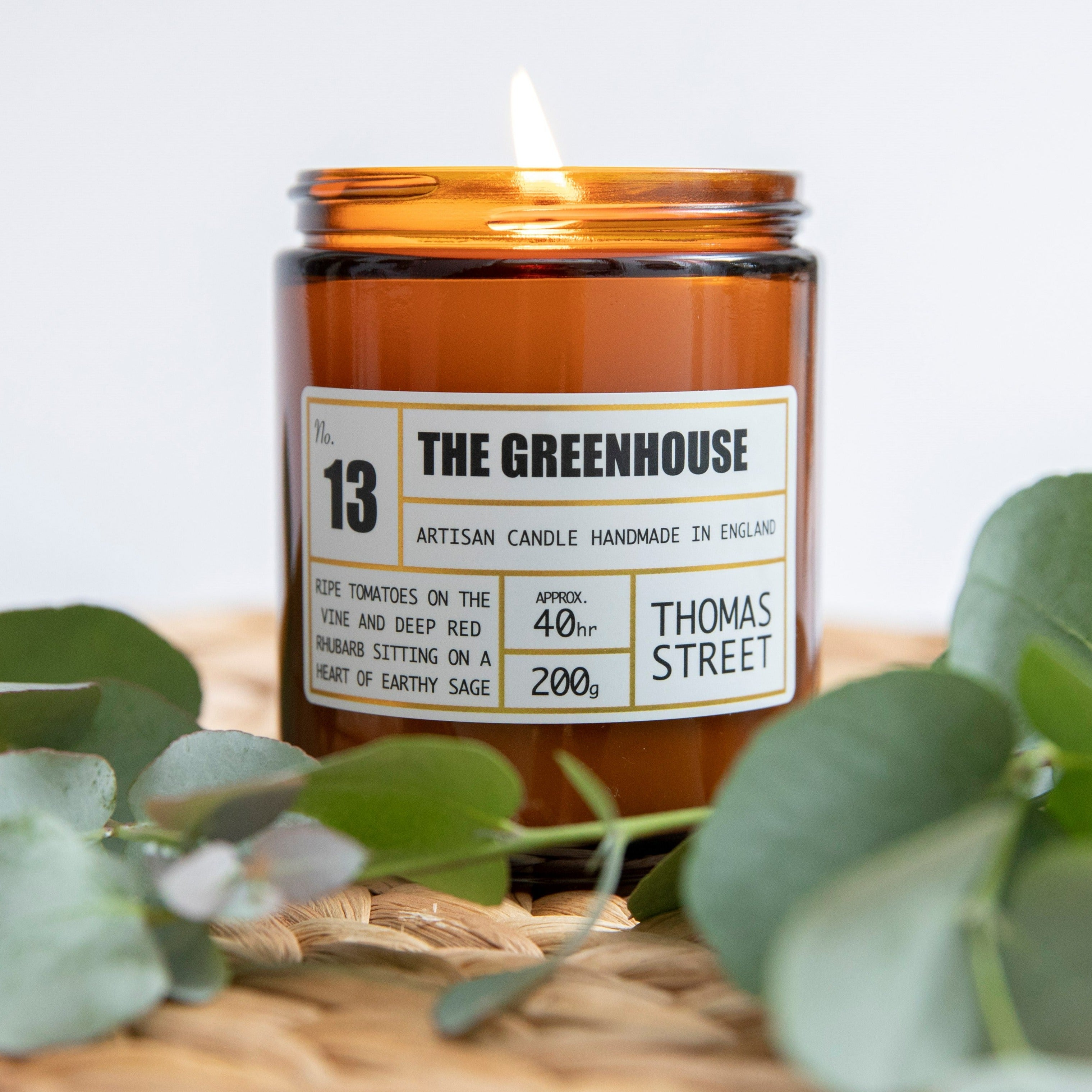 A soy wax scented candle named 'THE GREENHOUSE' from Thomas Street Candle Company, placed on a wicker surface with green leaves around it.