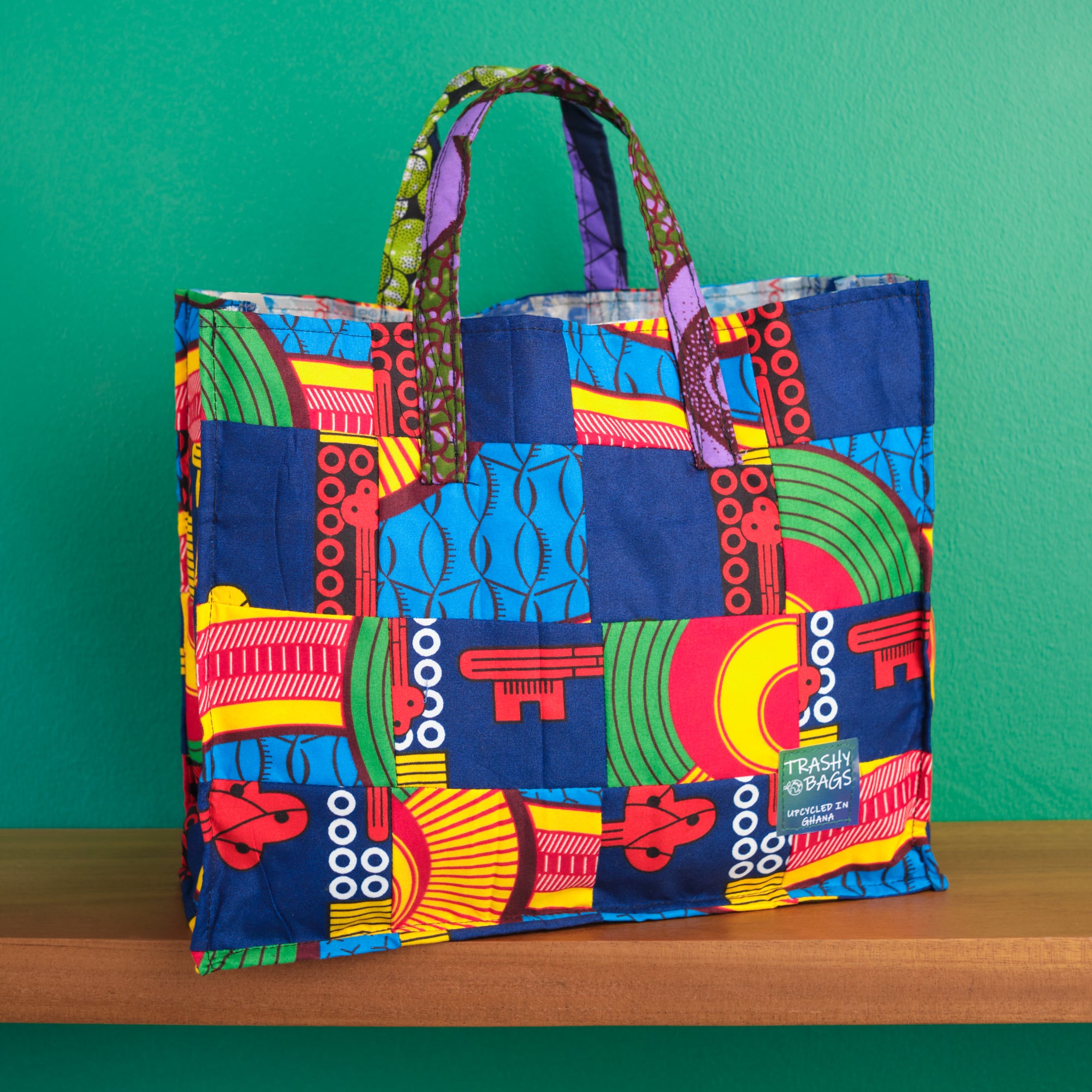 A multicolored recycled fabric tote shopper bag with a traditional African wax fabric print, standing on a shelf against a green background.