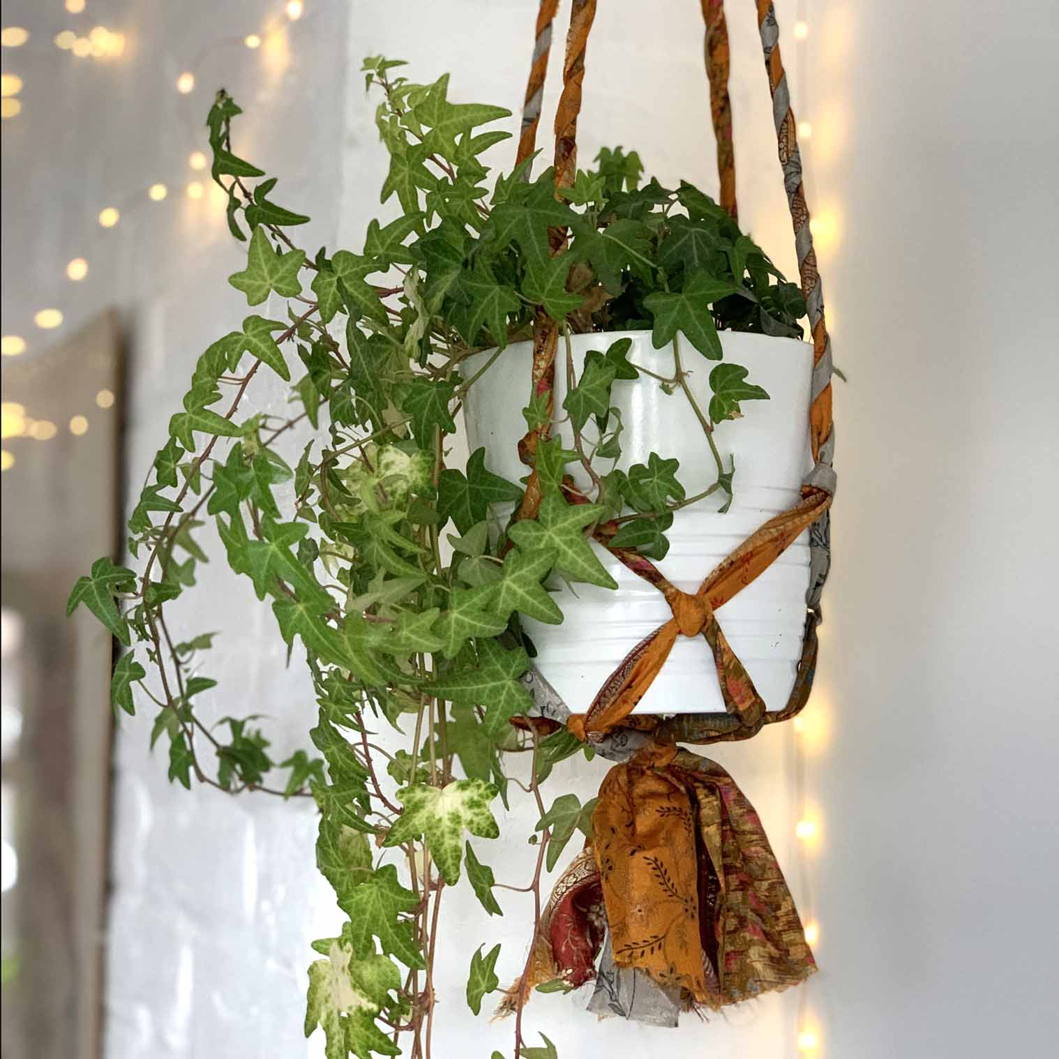 A plant hanger made from recycled sari material, with a metal ring for hanging, displayed with greenery in a white pot.