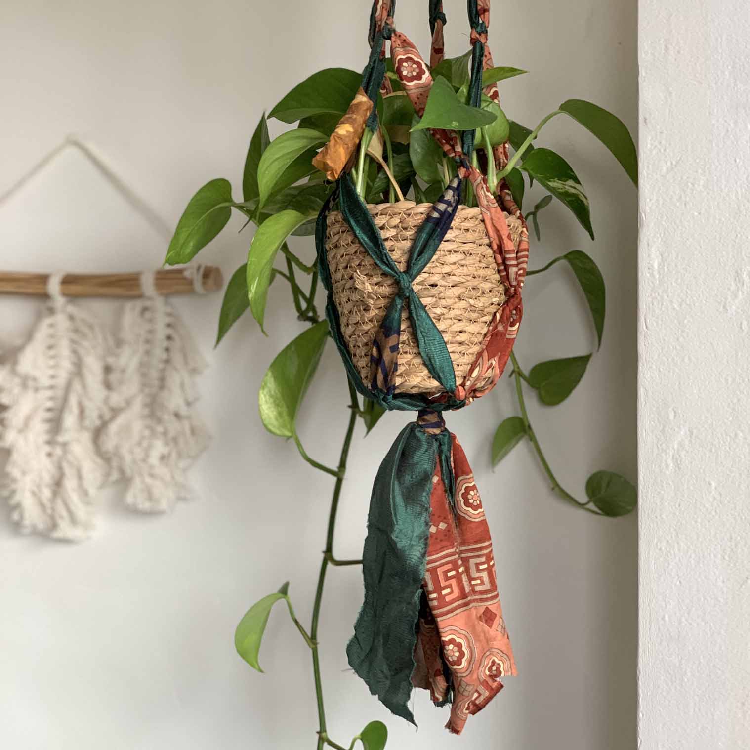 A plant hanger made from recycled sari fabric in various colors, hanging from a metal ring with a potted plant inside.