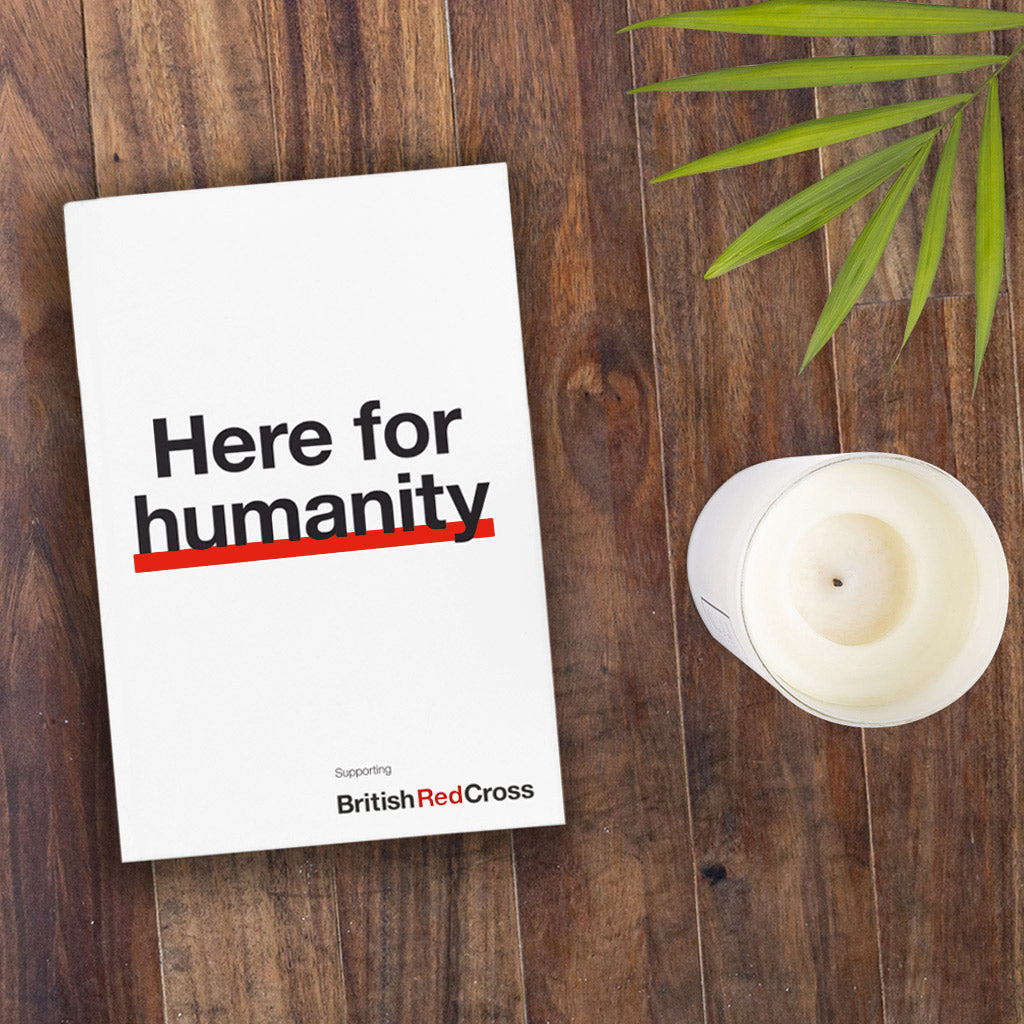 Book titled 'Here for humanity' by British Red Cross on a wooden surface with a candle and plant leaves.