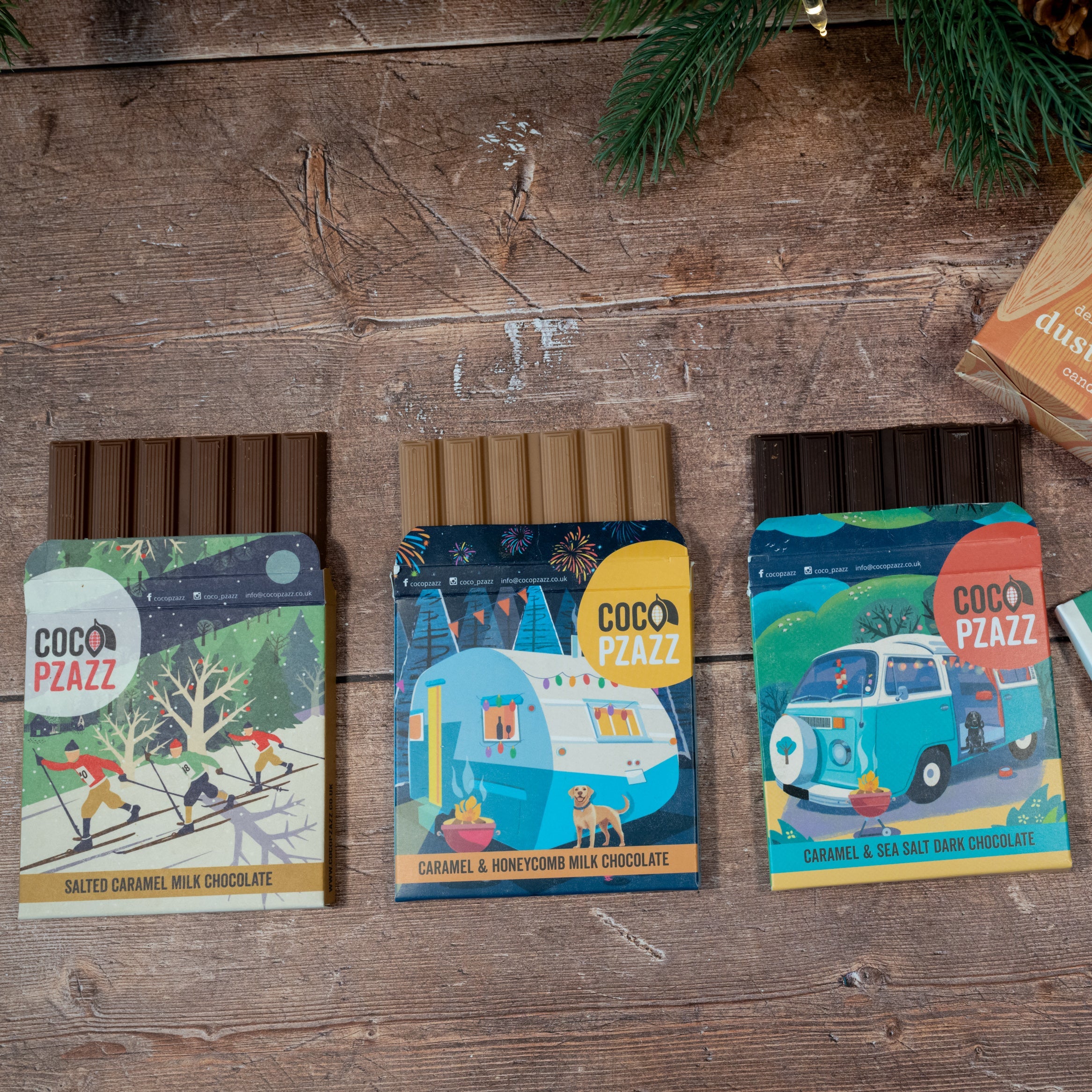Three packages of Coco Pazzz chocolate on a wooden surface with Christmas decorations.
