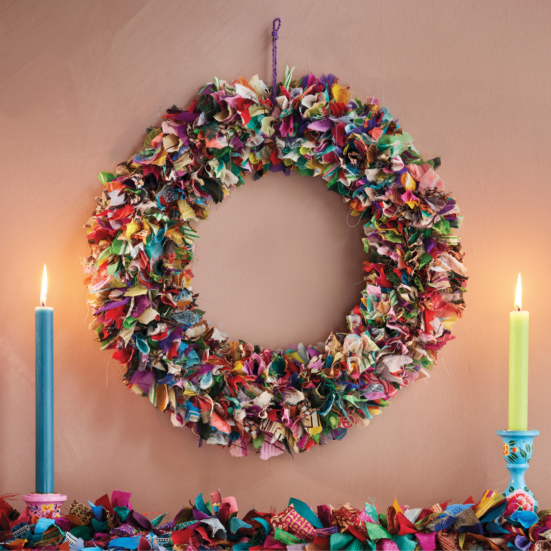 Colorful fabric wreath hanging on a wall with two candles below.